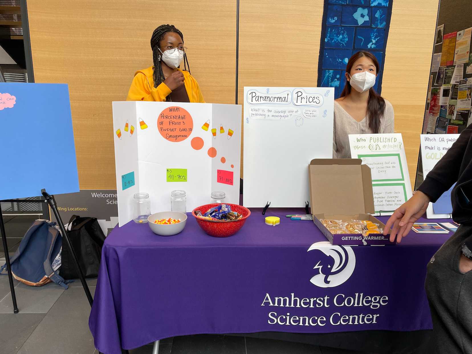 Candy and posters decorated with lettering and image cutouts sit on a table with a purple tablecloth and the words Amherst College Science Center on it. A Black woman and an Asian woman wearing masks stand people stand behind it.