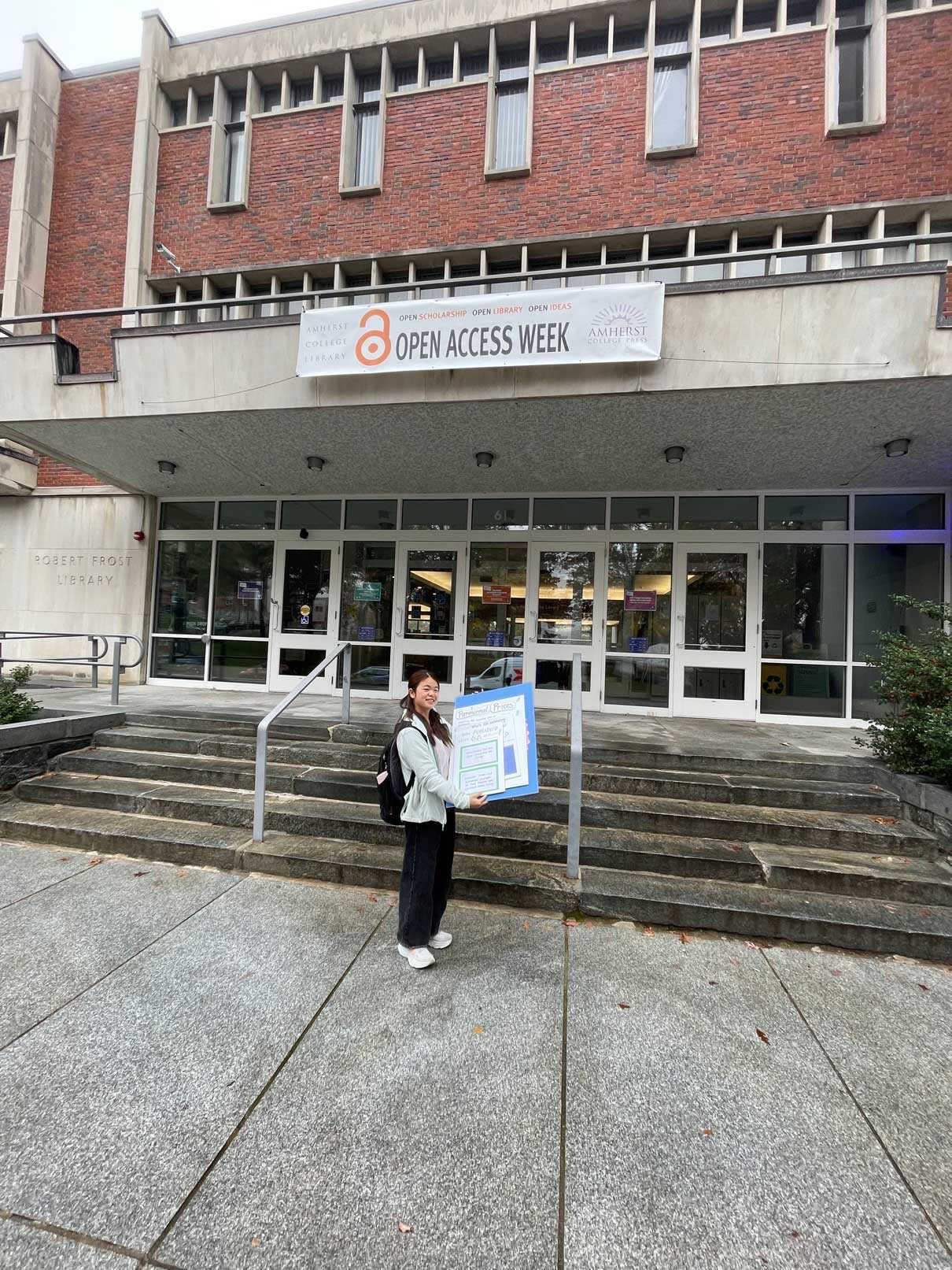 An Asian woman in white top and black pants holds posters in front of Robert Frost Library. A banner with the words Open Access Week hangs behind her.