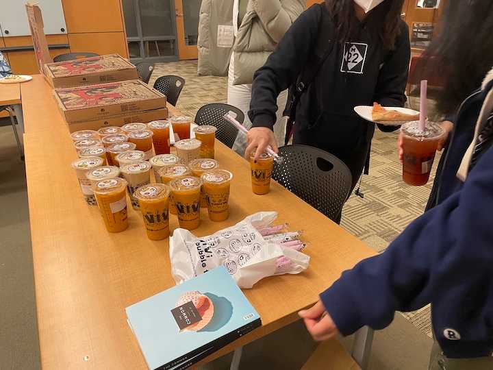 Boba teas and pizza boxes on a table. A person in a black sweatshirt is taking a tea. 
