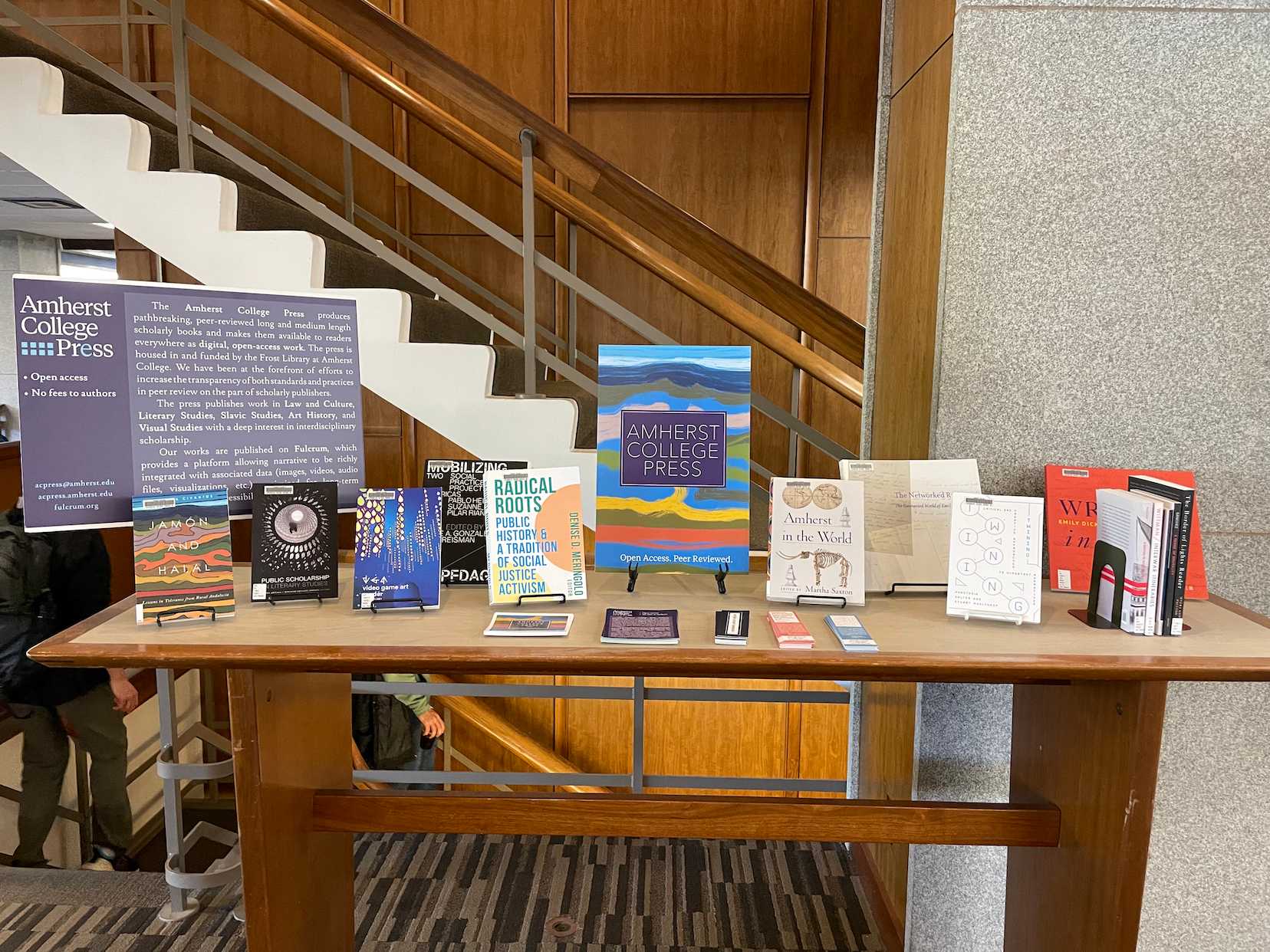 Books in stands and two posters with the words Amherst College Press sit on a table in front of stairs. 